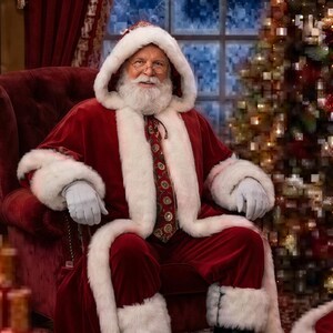 K&ouml;nnte beinhalten: Eine Weihnachtsmannfigur in einem tiefroten Samtmantel mit Kapuze, dickem wei&szlig;em Kunstfellbesatz, einer gemusterten Krawatte und hellgrauen Handschuhen. Die Figur tr&auml;gt einen wei&szlig;en Bart, eine Brille und schwarze Stiefel auf einem roten Sessel.