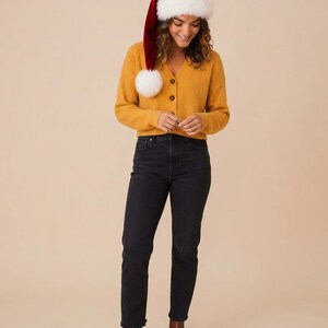 May include: A mustard yellow ribbed V-neck cardigan with buttons, paired with black denim jeans and brown leather ankle boots. A long red velvet Santa hat with a white faux fur trim and a large white pom-pom rests on the head.