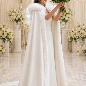 May include: A floor-length white cape featuring a thick, plush white faux fur trim along the hood and front edges, worn over a simple white gown. The garment is paired with a vibrant bouquet of deep red roses with green stems and leaves.
