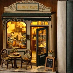 Puede incluir: Modelo en miniatura de un café, con el texto "Sunny Cafe" sobre la entrada. El modelo incluye una pequeña mesa y sillas, una pizarra y una puerta abierta. El interior está iluminado con una iluminación cálida, creando un ambiente acogedor.