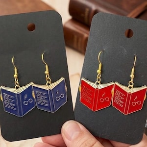May include: Two pairs of dangle earrings featuring open book charms. One pair is dark blue with gold-tone wire hooks, and the other is bright red with gold-tone wire hooks. Each book charm displays a lightning bolt and round glasses icon on the cover.