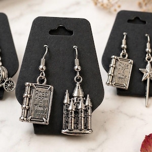 May include: Silver-toned earrings with a fairy tale theme. One pair features a castle design, while another displays a book with the words "Once Upon A Time." The earrings hang from silver hooks.