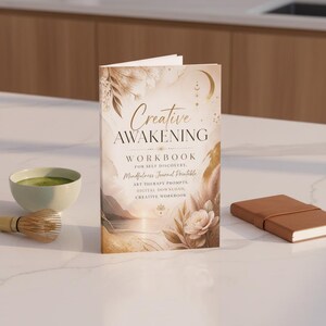 May include: A hardback workbook titled "Creative Awakening" with gold and beige floral and landscape illustrations. The book is open, displaying the title and subtitle, "Workbook for Self Discovery." A matcha bowl and a small brown notebook are also visible.