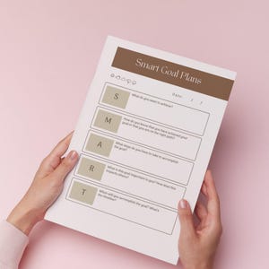 A person's hands are holding a sheet of paper titled "Smart Goal Plans" against a pink background.