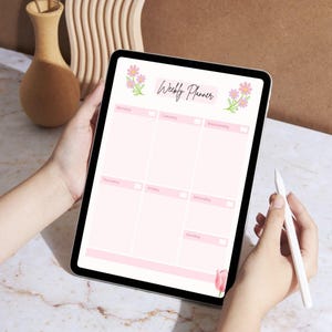 A person's hands holding a tablet displaying a pink and white weekly planner with a floral design.