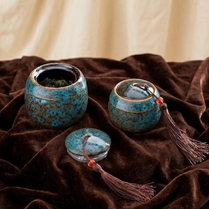 Puede incluir: Tres botes de té de cerámica con tapas, con un esmalte turquesa moteado. Un bote está abierto, revelando hojas de té. Cada bote tiene una borla con una pequeña cuenta roja. Los botes descansan sobre una superficie de terciopelo marrón oscuro.