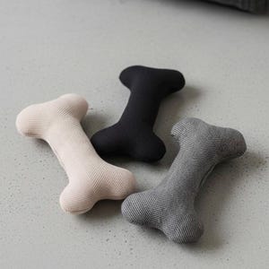 Peut inclure: Trois jouets pour chien en forme d'os, beige, noir et gris. Les jouets sont faits d'un tissu texturé et sont disposés sur une surface gris clair. Un panier pour chien est visible en arrière-plan.