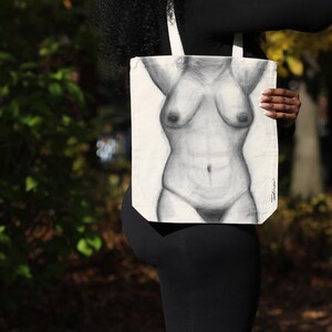 May include: A white tote bag featuring a detailed pencil drawing of a torso. The artwork depicts the front of a human body, with shading to define the form. The bag has white straps and is held against a background of green foliage.
