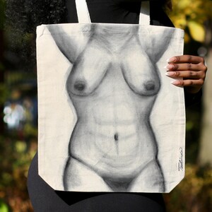May include: A beige tote bag featuring a grayscale pencil drawing of a human torso. The artwork depicts the front of a body with defined abdominal muscles and breasts. The bag has white straps and a signature in the bottom right corner.