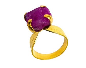 May include: A gold-colored ring featuring a large, raw, purple gemstone. The gemstone is held in place by four gold prongs. The ring band has a textured finish and a unique, open design.