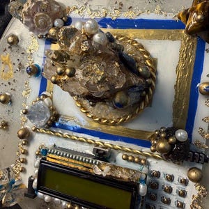 May include: A detailed, abstract art piece with a white base, featuring gold, blue, and clear accents. The design incorporates crystals, pearls, and a small electronic display. Gold leaf and metallic spheres add texture and visual interest to the artwork.