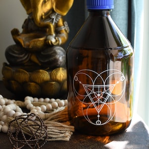 May include: A brown glass bottle with a blue cap and a white geometric design. A gold statue of Ganesha is in the background, along with a string of white beads and a decorative metal sphere.