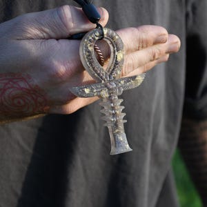May include: A clear ankh pendant with gold accents is held in a hand. The ankh, a symbol of life, features a loop, crossbar, and vertical element. The pendant is suspended from a black cord. The background is blurred, with a glimpse of green.