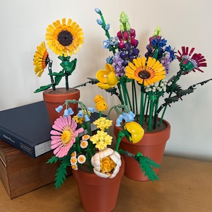 May include: A collection of colorful artificial flowers in terracotta pots. The arrangement includes sunflowers, lupines, and other blooms in shades of yellow, pink, purple, and blue. The flowers are displayed on a wooden surface.