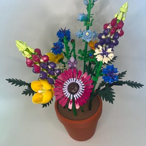 May include: A colorful bouquet of artificial flowers in a terracotta pot. The arrangement includes yellow, purple, blue, and white flowers with green stems and leaves. The flowers are arranged in a decorative display.