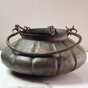 May include: A round, hammered copper pot with a wide, fluted body and a dark, weathered patina. The vessel features a thick, curved metal handle attached to the rim by two circular rings, showing signs of oxidation and age-related wear.