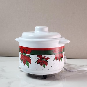 May include: A white ceramic electric pot with a lid, featuring a decorative band of red poinsettia flowers and green leaves against a white background with small black dots. The pot has two side handles, a white power cord, and black feet.