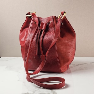May include: A deep red bucket bag with a textured leather surface, featuring a drawstring closure with matching leather cords, gold-toned metal hardware rings at the top, and a long, adjustable shoulder strap for carrying the bag.