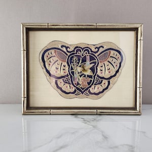 May include: Framed artwork featuring a detailed, symmetrical design in navy blue, gold, and pink. The artwork has a central floral and bird motif, set against a cream-colored background. The frame is a light, metallic gold color.