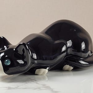 May include: A glossy black ceramic cat figurine in a crouching position. The cat features a white muzzle, white paws, and bright turquoise eyes. The smooth, reflective surface of the ceramic highlights the feline's curved, alert posture.