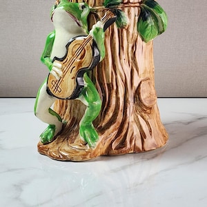 May include: A ceramic pencil holder shaped like a tree stump with a green frog playing a brown violin attached to the front. The tree stump has a textured brown bark finish with green leaves at the top, while the frog has a light cream belly.