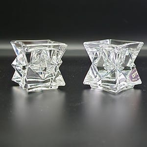 May include: Two clear, faceted crystal candle holders. Each has a square top and a geometric design. The candle holders are on a dark surface. One has a sticker that says "CRYS MADE IN".