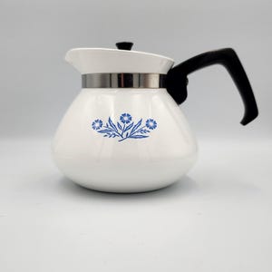 May include: A white teapot with a rounded body and a black handle. The teapot features a blue floral pattern on the front, a silver-colored metal band around the neck, and a black lid with a small knob. The surface is smooth and glossy.
