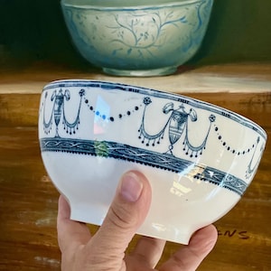 May include: A white ceramic bowl featuring a blue transferware pattern of neoclassical urns, draped swags, and beaded garlands along the rim, with a decorative braided border band encircling the upper section of the bowl's exterior surface.