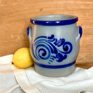 May include: A gray ceramic pot with a blue rim and handles, adorned with a blue floral design. A yellow lemon rests beside the pot on a white cloth, set against a wooden background.