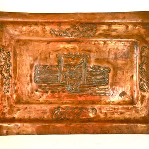 May include: A rectangular copper tray with a detailed etched design. The tray features a central scene of a structure with a figure, possibly a person or animal, and decorative floral elements on the corners and sides.