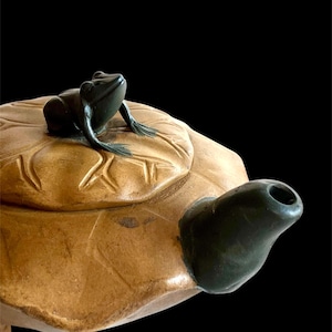 May include: A ceramic teapot with a frog-shaped spout and a frog figurine on the lid. The teapot is tan with carved leaf details, and the frog is dark green. The background is black.