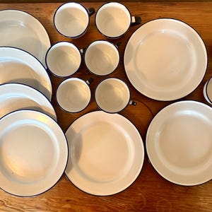 May include: A collection of white enamelware dishes and cups with a dark blue trim. The set includes plates of various sizes and several cups with black handles. The items are arranged on a wooden surface, creating a classic, vintage aesthetic.