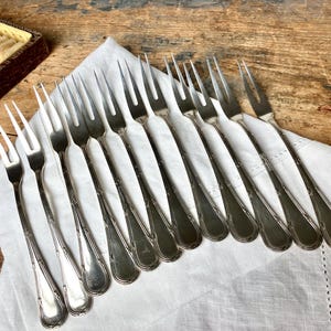 May include: A collection of silver forks arranged on a white cloth. The forks have a decorative design on the handles and are displayed on a wooden surface. The image suggests a vintage or antique silverware set.