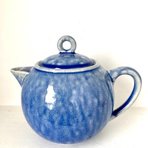 May include: A blue ceramic teapot with a white glaze and a round body. The teapot has a small handle and a lid with a ring on top.