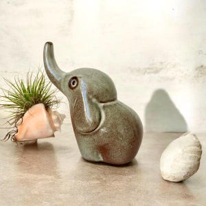 May include: A glossy, sage-green ceramic elephant figurine with a raised trunk and dark eye detail, positioned next to a light-colored seashell containing a spiky air plant and a separate, smaller, textured white seashell on a flat surface.