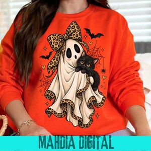 Peut inclure: Un sweat-shirt orange vif orné d'un motif représentant un fantôme blanc portant un grand nœud à imprimé léopard et tenant un chat noir. Le dessin comprend des chauves-souris noires, des toiles d'araignée et des petits cœurs orange.