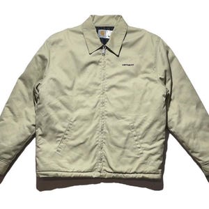 May include: A light tan Carhartt jacket with a black collar and a zipper closure. The jacket has two front pockets and the Carhartt logo embroidered on the chest. The jacket is made of a durable material and is suitable for various weather conditions.