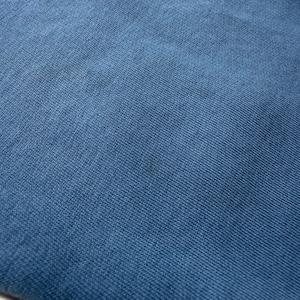 May include: Close-up of a textured, blue fabric. The material appears to be a knit or woven textile, with a subtle pattern visible. The color is a muted, medium blue, with slight variations in tone across the surface.