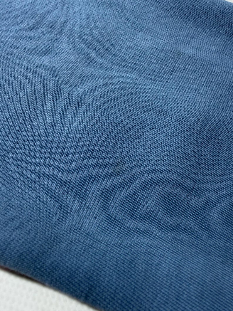 May include: Close-up of a textured, blue fabric. The material appears to be a knit or woven textile, with a subtle pattern visible. The color is a muted, medium blue, with slight variations in tone across the surface.