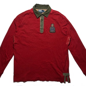 以下が含まれることがあります： 緑の襟とボタン留めの前立てが付いた、長袖の赤いシャツ。胸に紋章パッチ、袖にグリーンのアクセントが付いています。クラシックなスタイルです。