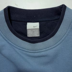 May include: Close-up of a blue sweatshirt with a navy blue collar and trim. The Nike logo is visible on a tag inside the collar. The sweatshirt has a ribbed texture around the collar.
