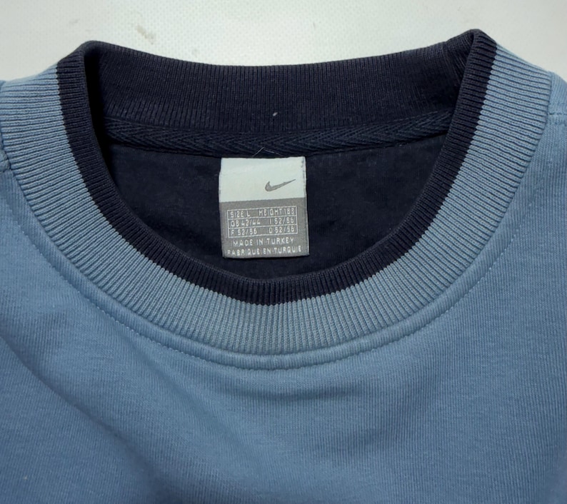 May include: Close-up of a blue sweatshirt with a navy blue collar and trim. The Nike logo is visible on a tag inside the collar. The sweatshirt has a ribbed texture around the collar.