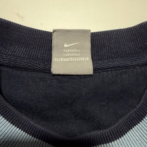 May include: Close-up of a navy blue crew neck sweatshirt with a light blue stripe at the neckline. A gray tag with the Nike logo and text is sewn into the collar.