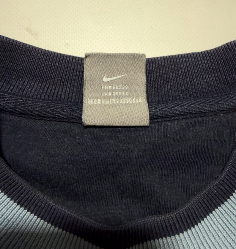 May include: Close-up of a navy blue crew neck sweatshirt with a light blue stripe at the neckline. A gray tag with the Nike logo and text is sewn into the collar.
