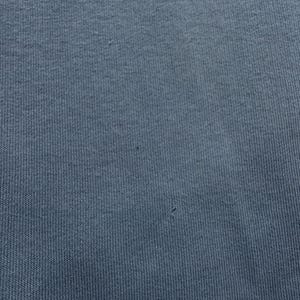 May include: Close-up of a textured, blue-grey fabric. The material has a ribbed pattern, with subtle variations in color and texture. The fabric appears to be a knit or woven textile, with a soft, matte finish. The image shows the details of the textile.