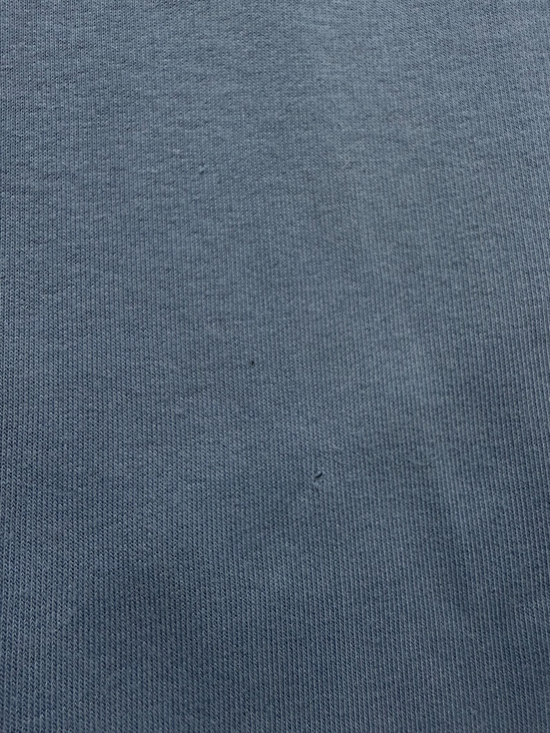 May include: Close-up of a textured, blue-grey fabric. The material has a ribbed pattern, with subtle variations in color and texture. The fabric appears to be a knit or woven textile, with a soft, matte finish. The image shows the details of the textile.