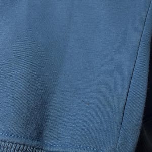 May include: Close-up of a blue sweatshirt. The fabric is a solid, muted blue color with a ribbed knit cuff at the bottom. The stitching is visible along the seams.
