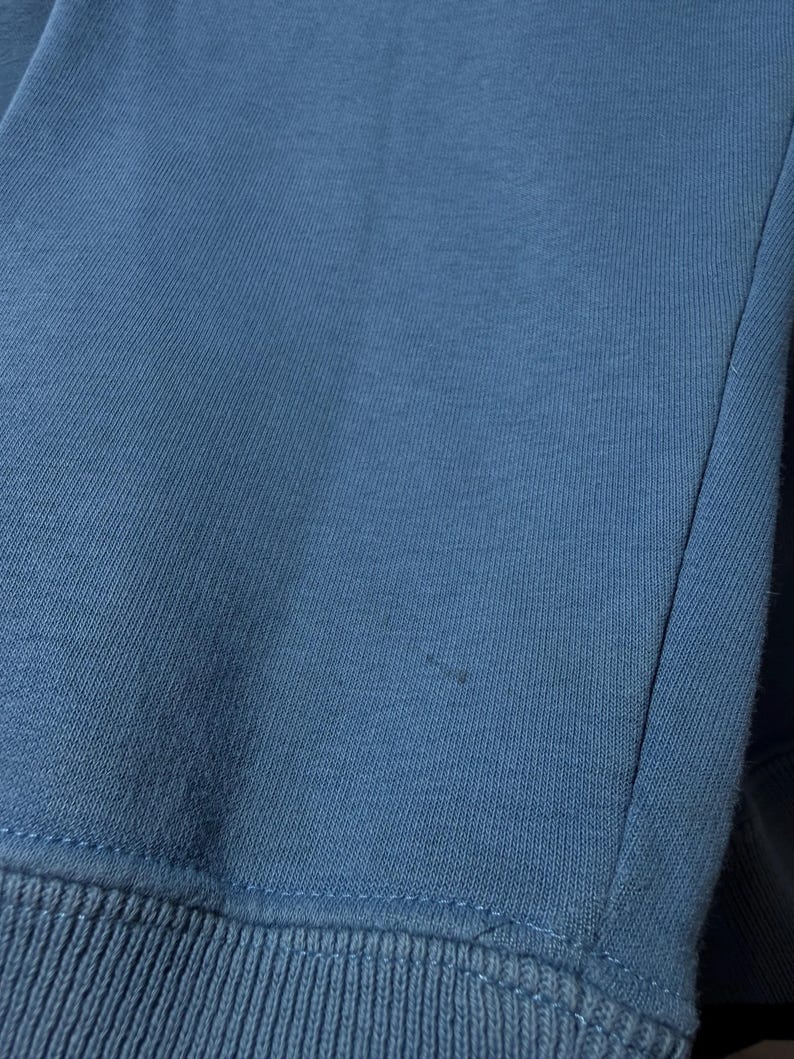 May include: Close-up of a blue sweatshirt. The fabric is a solid, muted blue color with a ribbed knit cuff at the bottom. The stitching is visible along the seams.