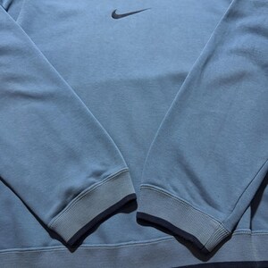 May include: A light blue sweatshirt with a dark blue collar, cuffs, and hem. The Nike swoosh logo is embroidered on the upper left chest. The long sleeves have ribbed cuffs. The sweatshirt is made of a soft, comfortable material.