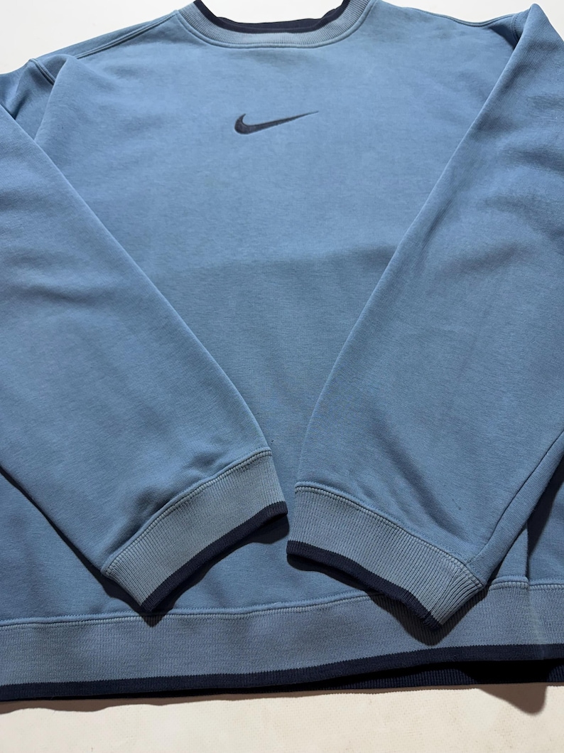 May include: A light blue sweatshirt with a dark blue collar, cuffs, and hem. The Nike swoosh logo is embroidered on the upper left chest. The long sleeves have ribbed cuffs. The sweatshirt is made of a soft, comfortable material.